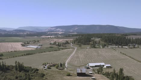 aerial-fly-out-industrial-farm-land-bare-mountains-within-a-valley-with-curvy-roads-tall-pine-trees,-dairy-wheat-cattle-barns-on-the-countryside-in-British-Columbia-off-the-freeway-to-Alberta-CA-3-5