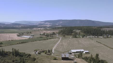 Vuelo-Aéreo-Fuera-De-Tierras-Agrícolas-Industriales-Montañas-Desnudas-Dentro-De-Un-Valle-Con-Carreteras-Con-Curvas-Pinos-Altos,-Graneros-De-Ganado-De-Trigo-Lechero-En-El-Campo-En-Columbia-Británica-Junto-A-La-Autopista-A-Alberta-Ca-3-5