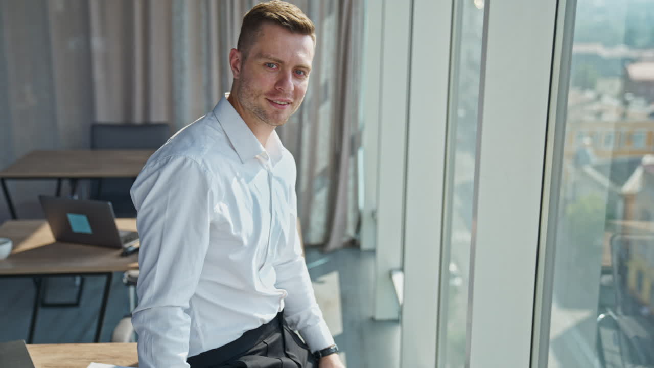 Calm confident young businessman in white shirt standing at the window. Man looks into sunlit window and then to the camera.