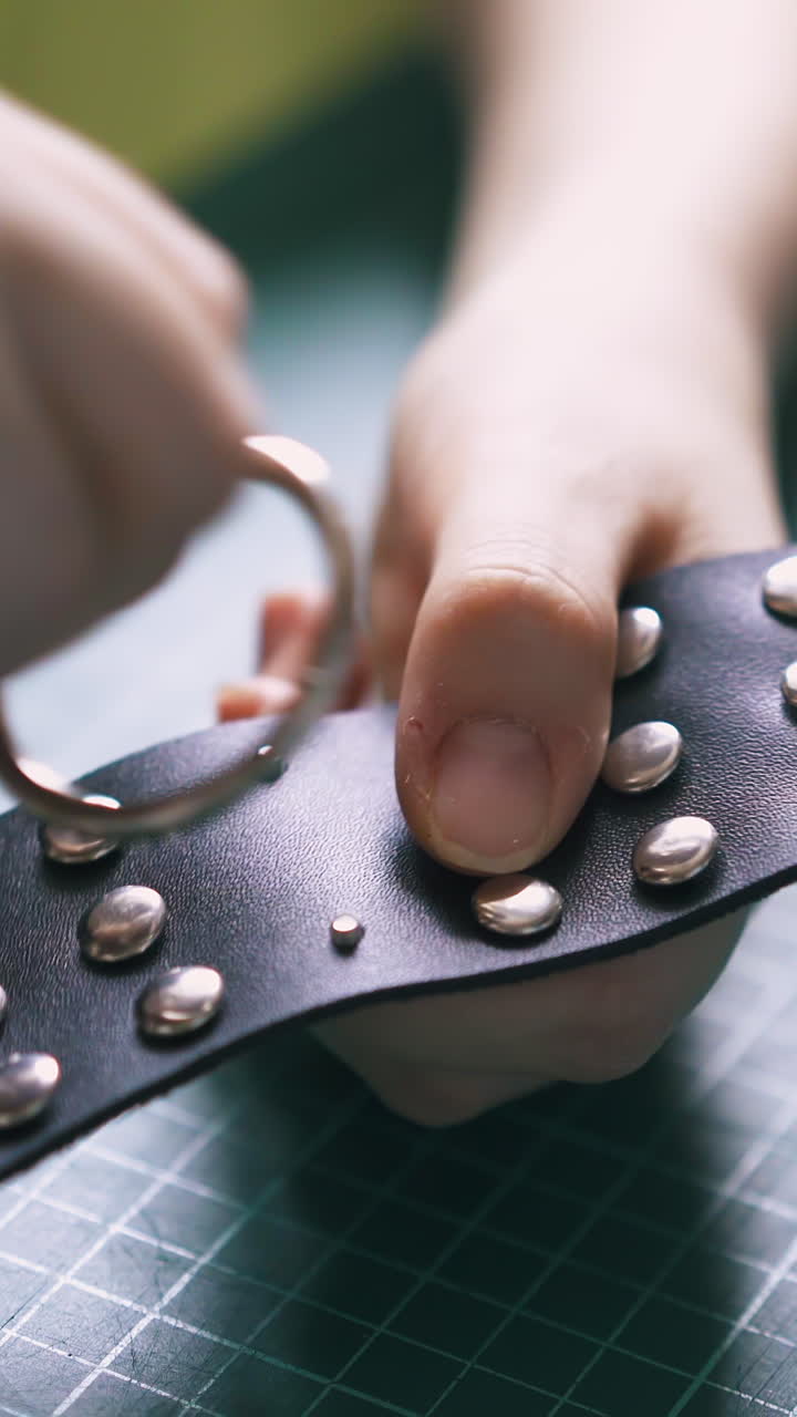 CU: Skilled girl designer inserts fashionable silver accessory into black leather choker with holnitens close view
