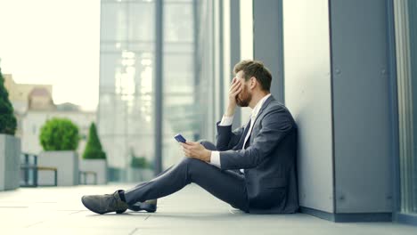 sad depressed entrepreneur in formal suit worker man sitting near outdoors street near modern office business center.