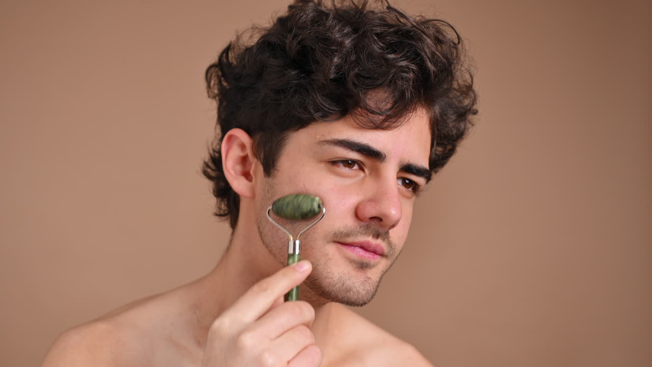 A young caucasian man with stubble beard is doing a facial massage using a roller