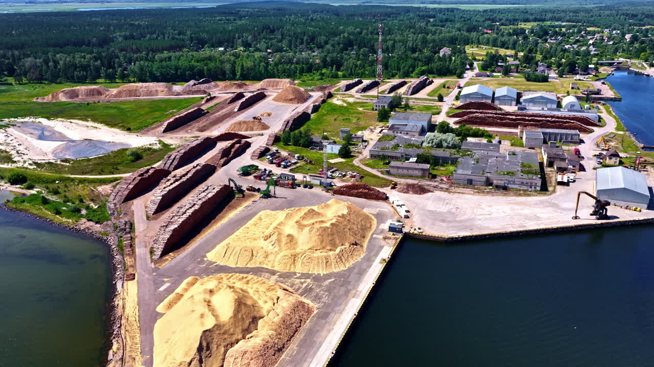 Aerial view of a coastal industrial yard filled with large piles of lumber and sawdust, machinery, storage rows, and a loading dock beside calm blue water