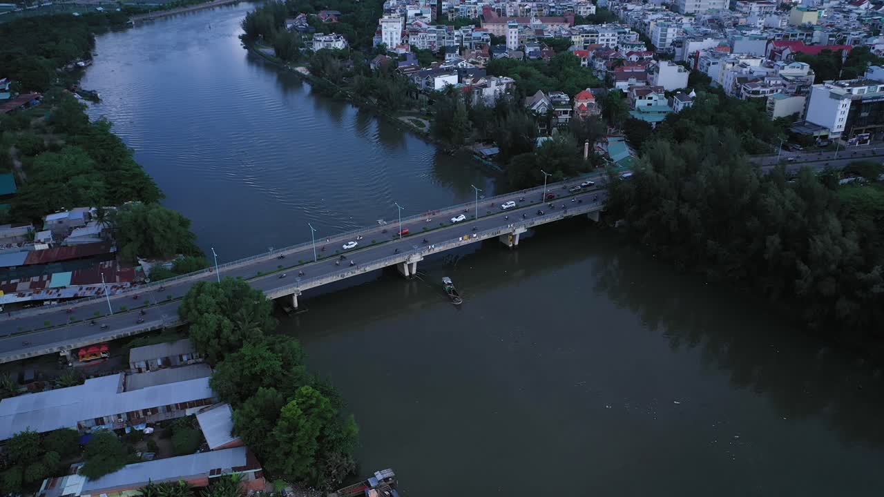 toma aérea de un barco fluvial que pasa por debajo de un puente de tráfico urbano y viviendas frente al mar a lo largo de un canal en la ciudad de ho chi minh, vietnam, a la luz de la tarde