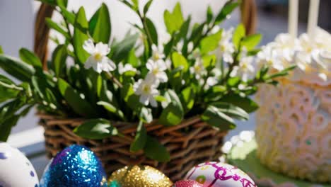 composition of shiny easter eggs in blue, gold, pink and white with glitter and decorations, next to a wicker basket with white flowers and green leaves, isolated on a white background