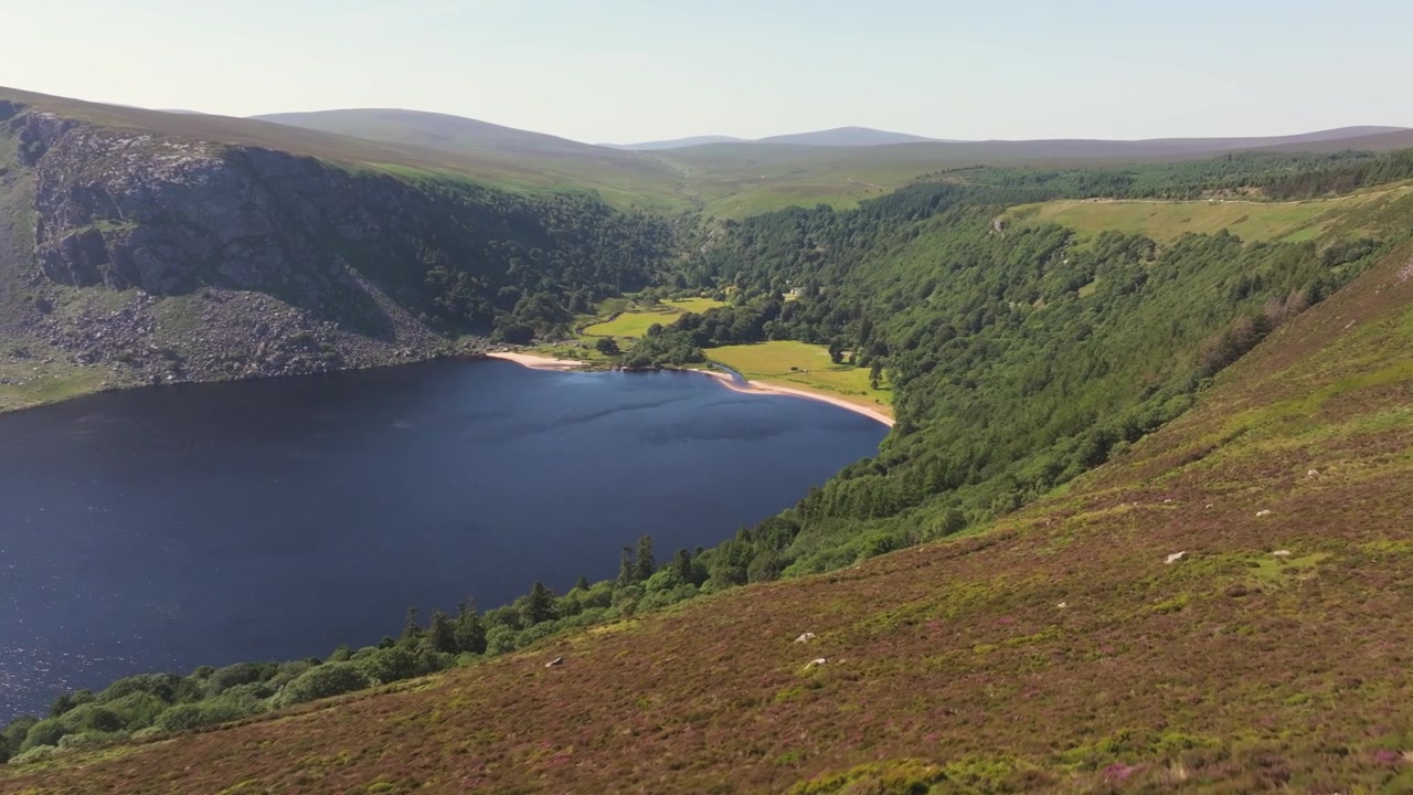 Guinness Lake bukter seg under lyngkledde åser og en skogkledd dal mens luftfotoet beveger seg over strandlinjen på en klar dag