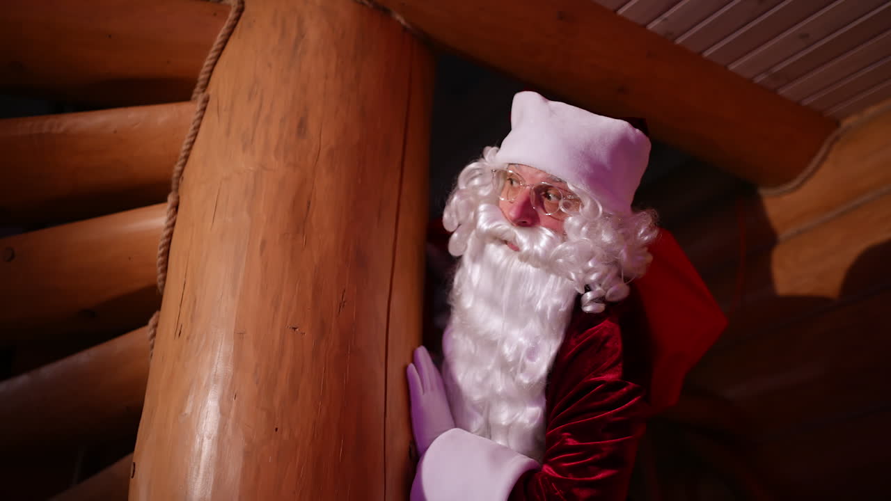 Santa Claus hiding in a wooden house. Authentic Santa with white beard and red costume with a sack with presents looking out of wooden pillar.