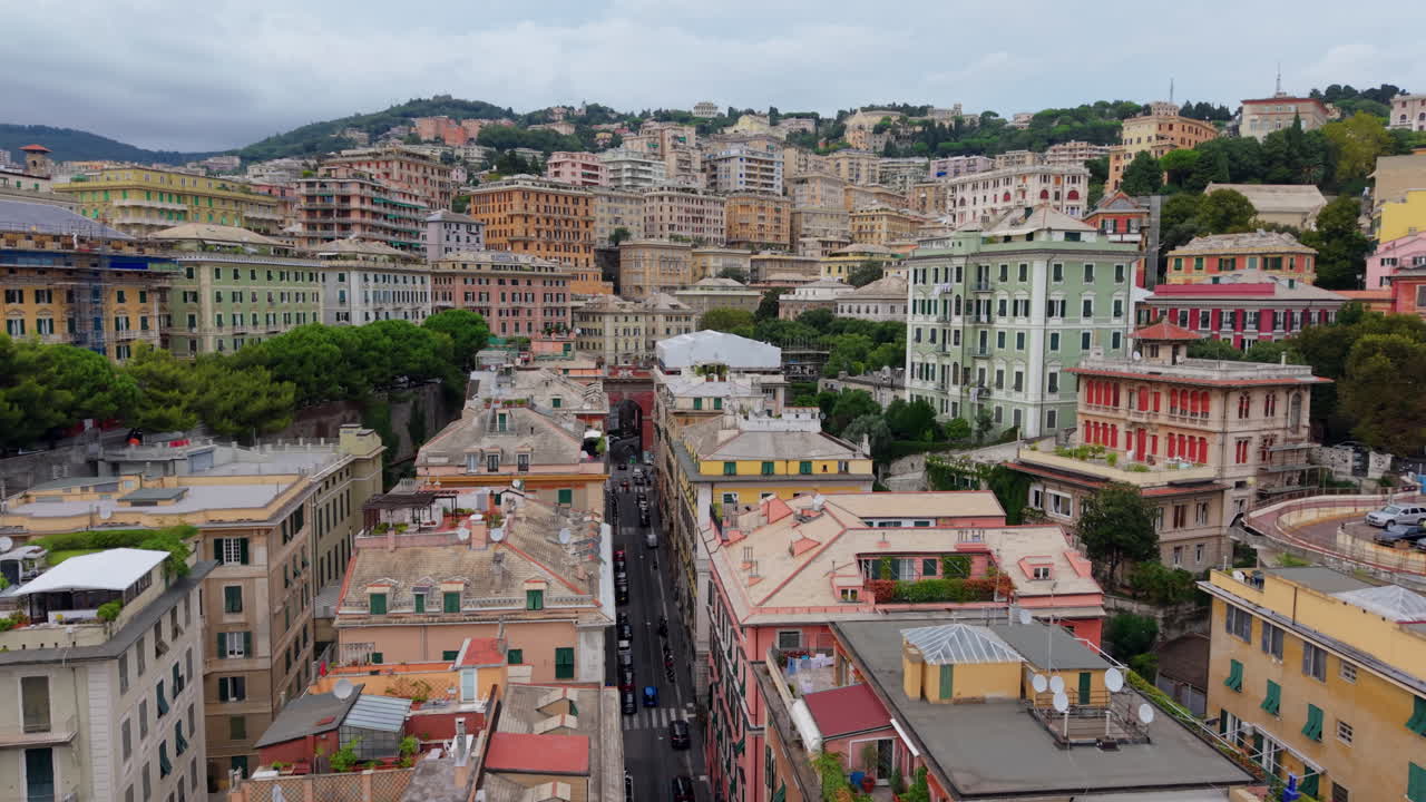 Drone flies forward at low altitude above the colorful rooftops of Genoa, Italy, following a street with moving vehicles and showing the hillside cityscape in the background