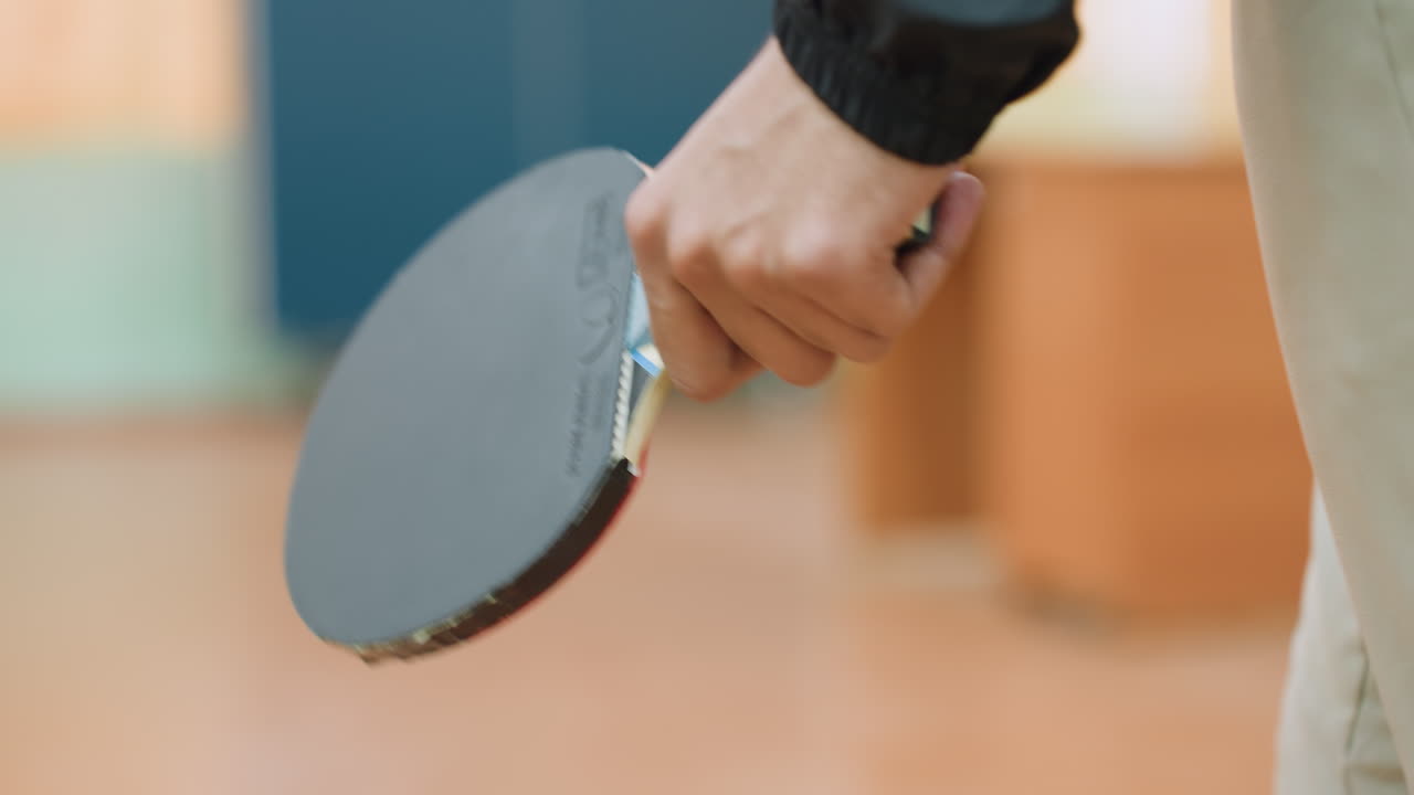 Close up of athlete holding racket firmly while walking toward tennis table with blurred indoor background, showing preparation and focus before starting sports activity