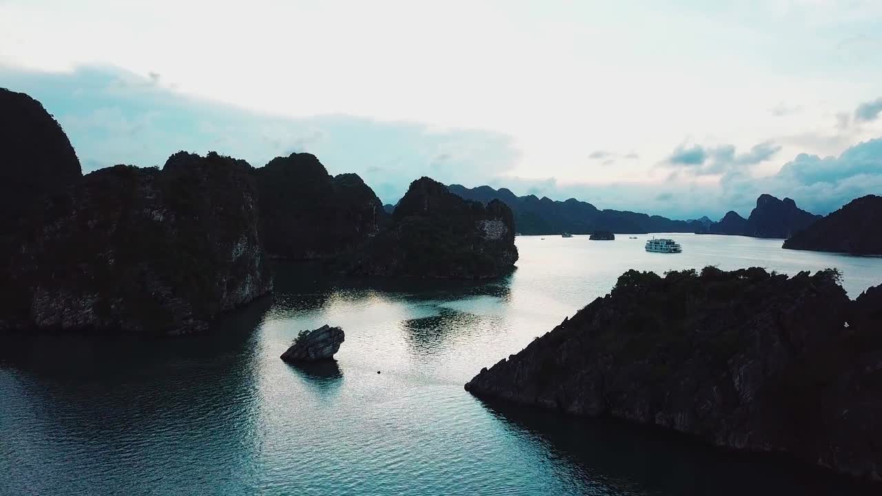 Aerial view of calm bay at golden hour with fishing boat drifting between dramatic limestone islands. Soft evening light reflects on tranquil water, highlighting natural seascape beauty