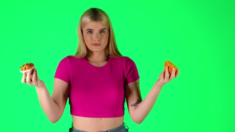 joven rubia con pastel y rebanada de naranja, elección entre comida saludable y no saludable, fondo verde tiro de estudio
