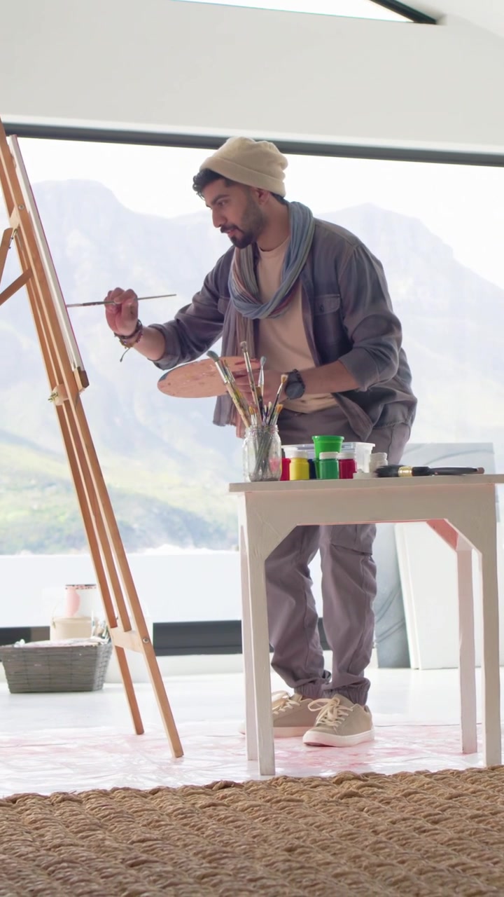 Vertical video: Picking up brush, Indian artist painting at studio, wearing beanie holding palette