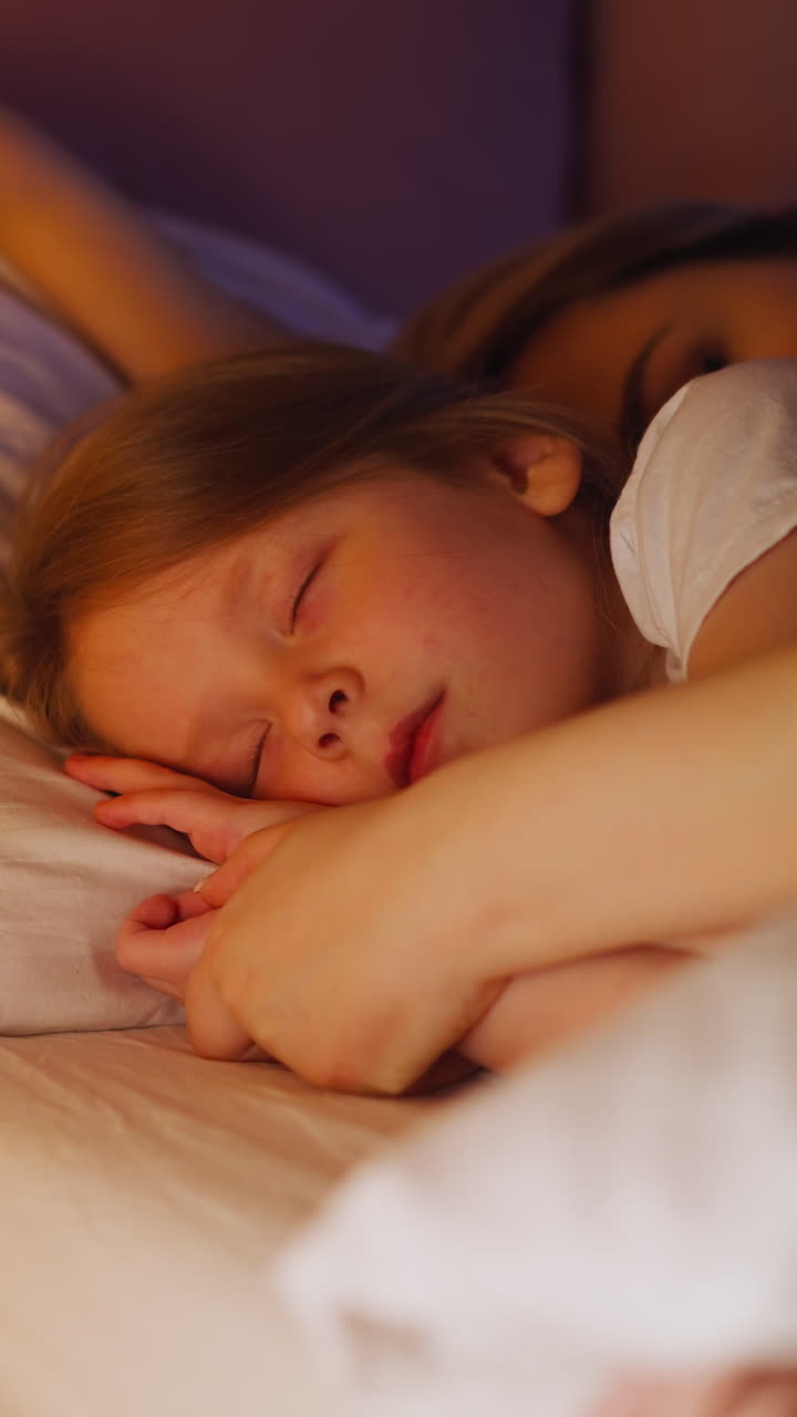 Sleepy mother embraces blonde little daughter lying together in bed with large pillows and warm blanket in dimly lit bedroom closeup slow motion