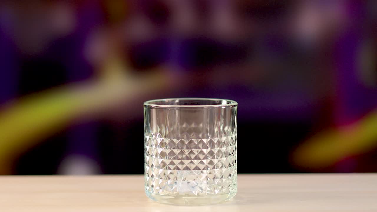 Ice cubes drop into a textured glass on a wooden surface against a colorful blurred background
