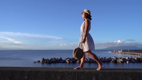 Chica-Japonesa-Con-Vestido-De-Verano-Sosteniendo-Una-Bolsa-Caminando-Frente-Al-Océano-En-Un-Hermoso-Día-Despejado---Amplia-Toma-De-Seguimiento-Lateral