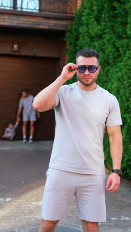sportive caucasian man puts on sunglasses. woman with a little boy stand holding hands at blurred backdrop. vertical video