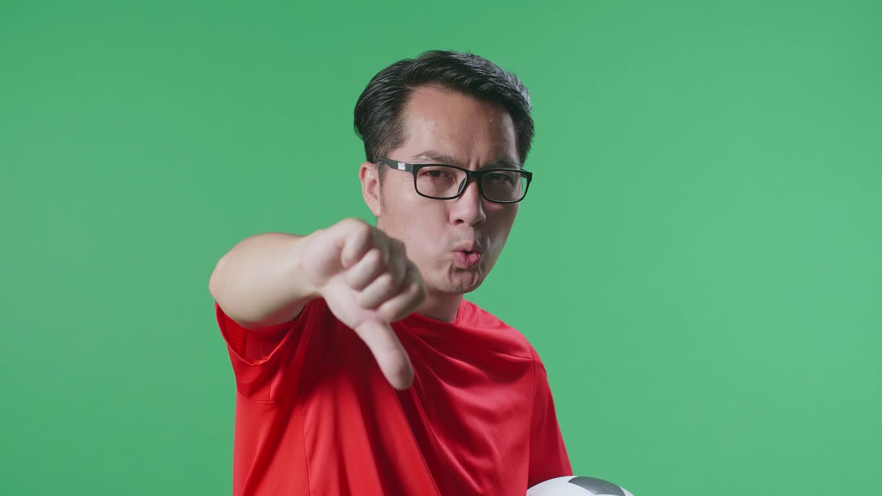 녹색 스크린 배경에서 축구를 응원하는 동안 패자를 조롱하는 몸짓으로 엄지손가락을 아래로 보여주는 공을 가진 아시아 남자의 클로즈업