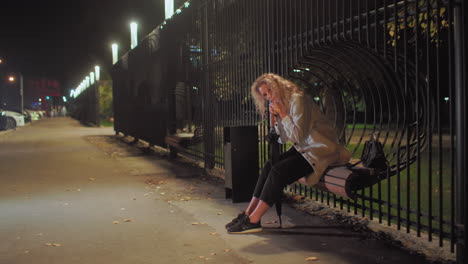 tired lady seated on decorative wooden bench attached to iron fence, resting head on umbrella with bag beside her, street lights shining, blurred group of people visible in distant background