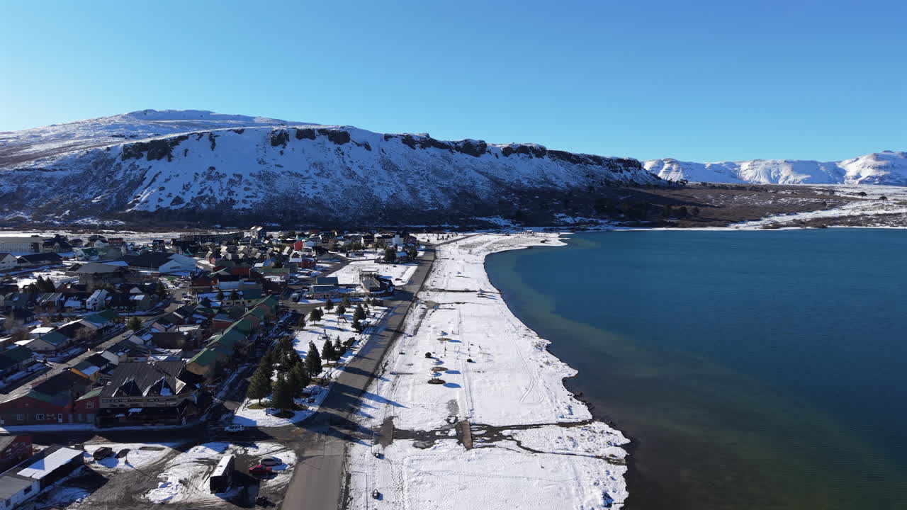 Drone panning shot from Caviahue lake toward the town, with snowy shoreline, houses, and sunlit mountains in Patagonia, Argentina