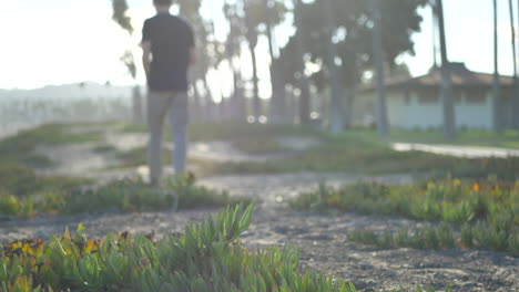 A-low-shot-of-a-young-man-and-his-feet-walking-out-of-focus-into-the-background-along-the-sandy-beach-and-palm-trees-in-Santa-Barbara,-California