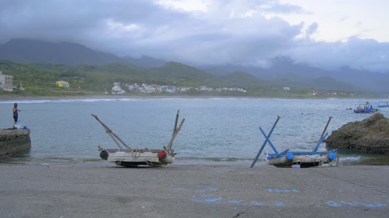 Fisherman and beached rafts at a boat ramp with mountains and cloudy sky in background, filmed in wide as establishing shot