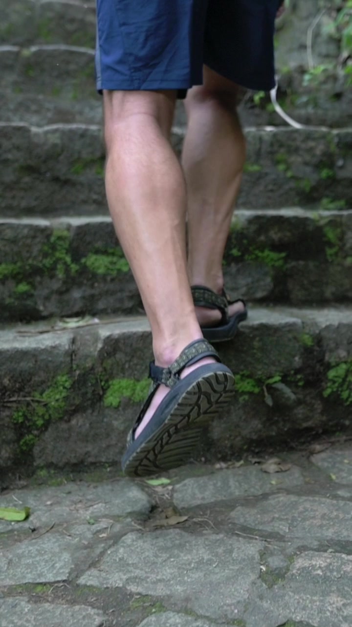 Schermo verticale. Primo piano di gambe che salgono gradini di pietra muschiosi, escursionista con sandali e pantaloncini, muscoli del polpaccio in tensione, passo lento e costante su un sentiero tropicale ombreggiato a Da Nang, superfici di pietra texturizzate.