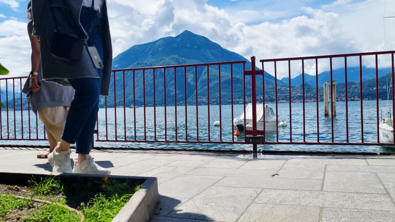 Close up of people walking along the promenade at Lake Como, with the lake, boats, and a mountain in the background on a partly cloudy day in Varenna Italy