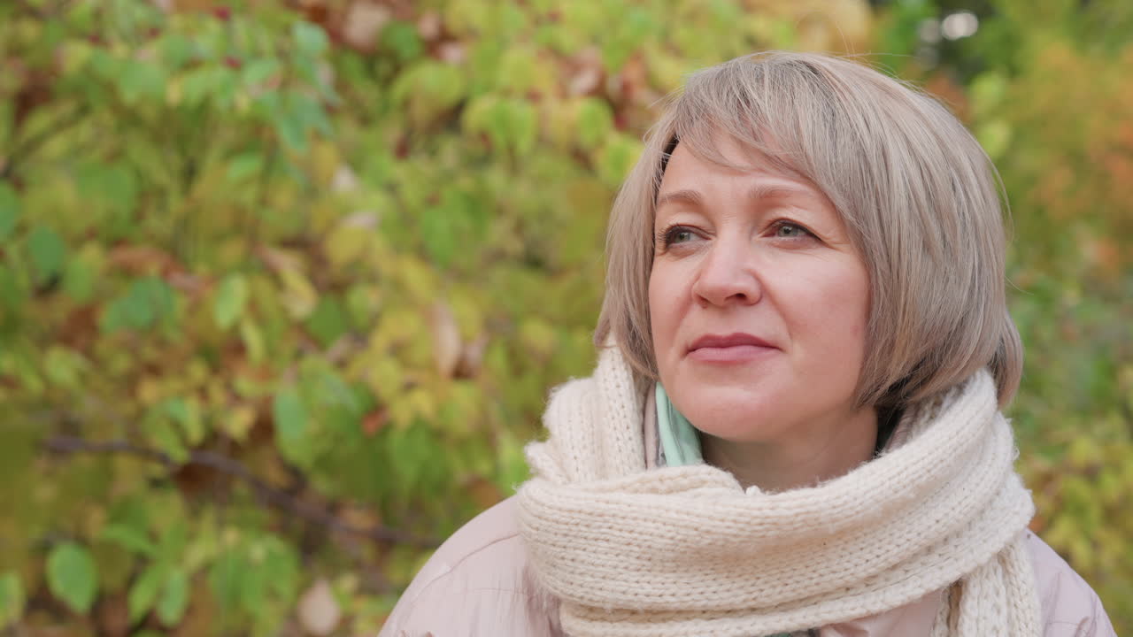 Close up of middle aged woman with short hair wearing soft knitted scarf and light jacket, smiling warmly while gazing at someone during peaceful walk in colorful autumn forest with golden foliage