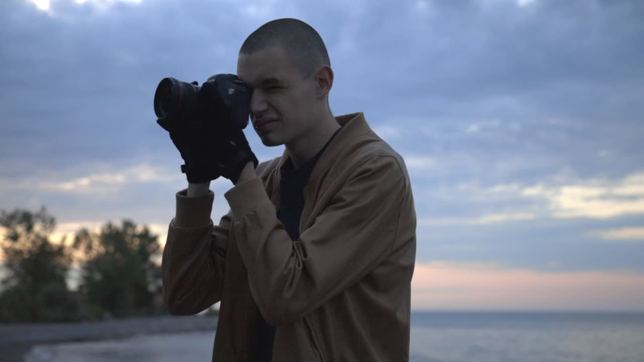 A Young Man Standing In The Shoreline While Taking Pictures Of The Area During Sunset - Close Up Shot