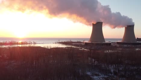 rising up aerial drone footage of nuclear power plant cooling towers at sunrise sunset with steam and smoke and bright sun winter