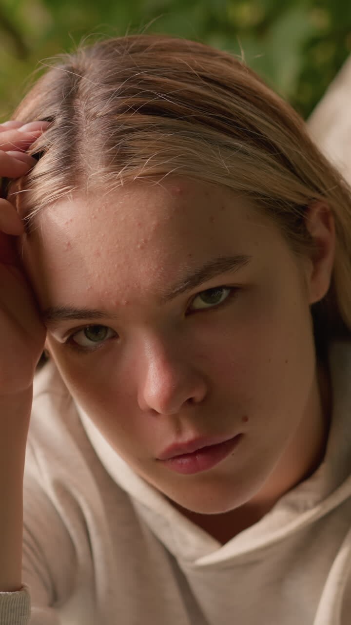 молодая женщина, сидящая на земле, опирается головой на левую руку, всматриваясь вверх с задумчивым выражением лица, на фоне которой изображена кора деревьев, листья и зелень