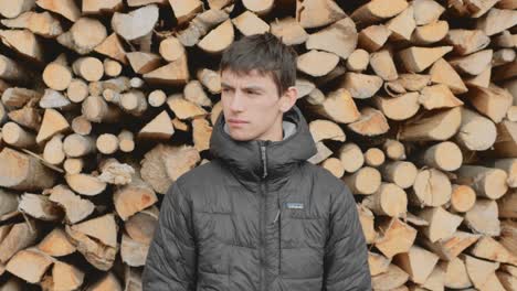 young man in black jacket standing in front of a big woodpile looking into the camera and turnng head