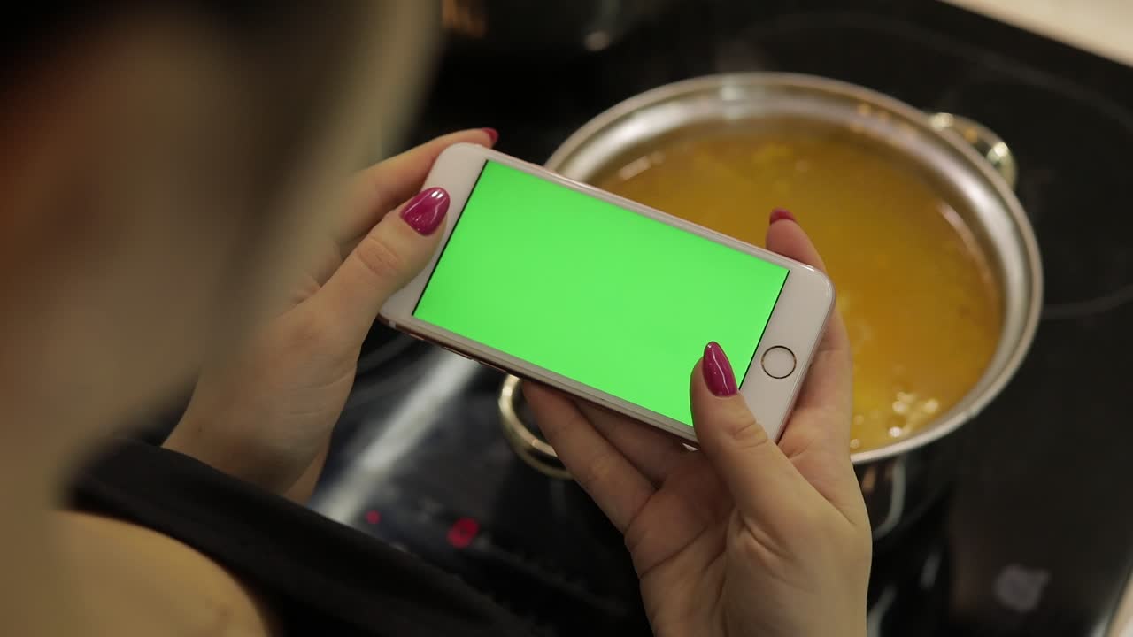 mujer está sosteniendo un teléfono inteligente con una pantalla verde, y cocinando sopa