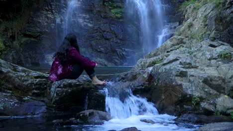 girl sitting on rock with waterfall flowing water from mountain at forest from flat angle video taken at thangsingh waterfall shillong meghalaya india