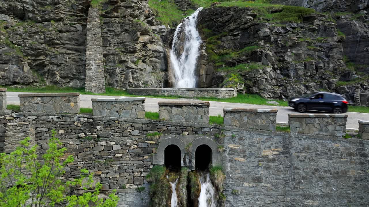 A scenic mountain waterfall cascading over rocks and through a historic stone structure with a road and passing car.