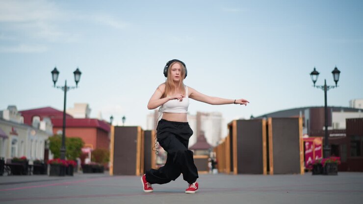 young woman dancing on street in empty city plaza wearing white top and loose black pants with red sneakers, spinning and stepping through expressive freestyle choreography under clear daytime sky