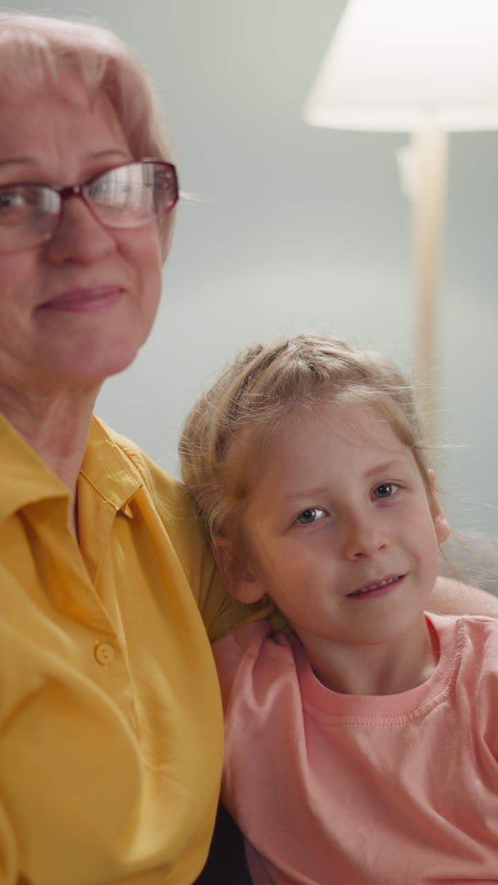 Smiling mature lady with glasses embraces loving little granddaughter. Family members pose sitting on couch in living room slow motion. Love and care in family