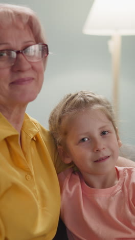 smiling mature lady with glasses embraces loving little granddaughter. family members pose sitting on couch in living room slow motion. love and care in family