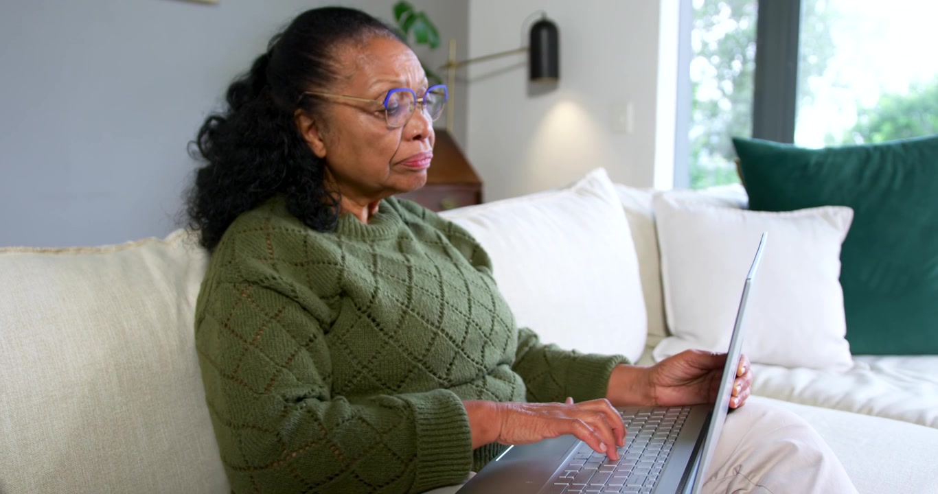 Senior African American woman in green sweater and glasses typing on laptop on sofa, checking work