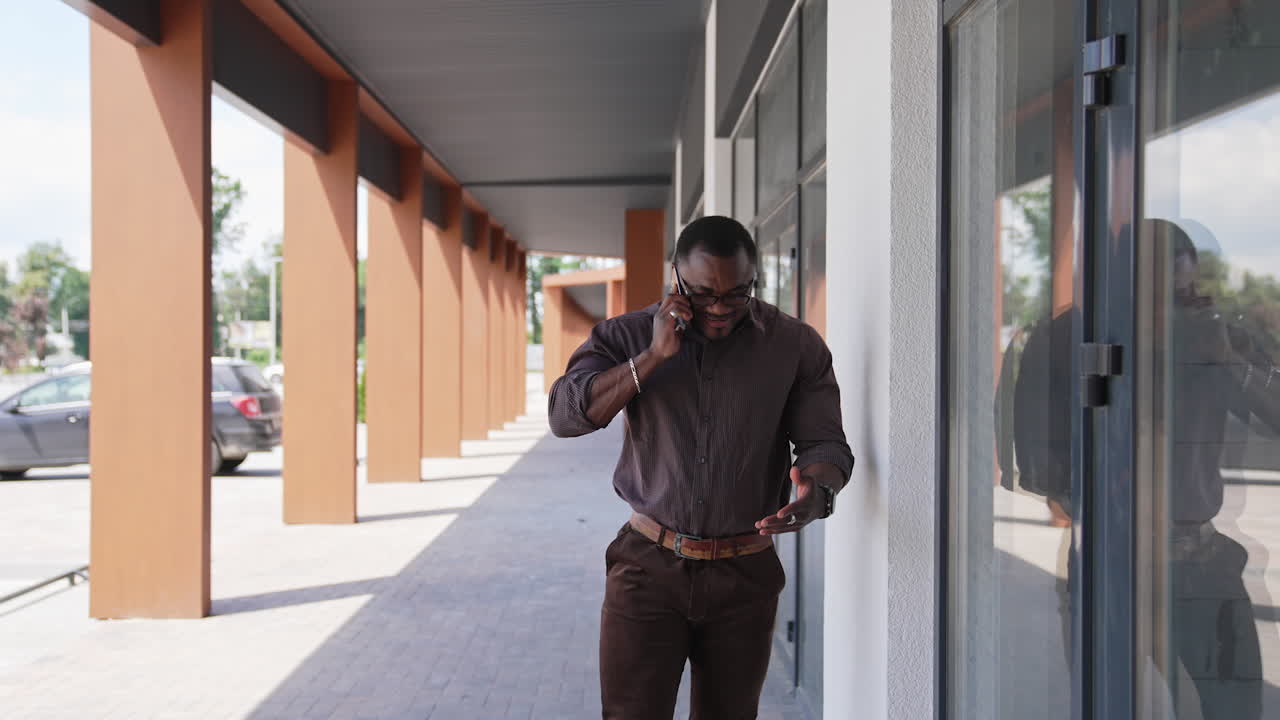 Afro american leader man having a phone talk outdoors. Athletic man with dark skin in shirt and pants talking the phone next to the modern building.