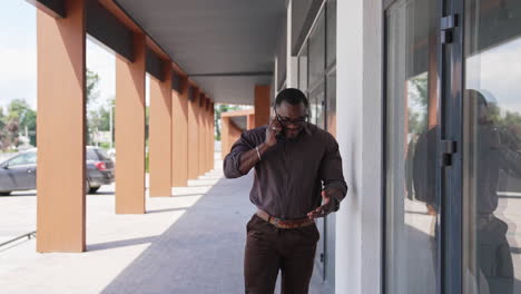 afro american leader man having a phone talk outdoors. athletic man with dark skin in shirt and pants talking the phone next to the modern building.