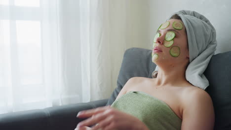 woman relaxing on couch wrapped in towel, folded hands together, cucumber slices placed on her face and lap, serene skincare and self-care moment, calm and rejuvenating spa experience