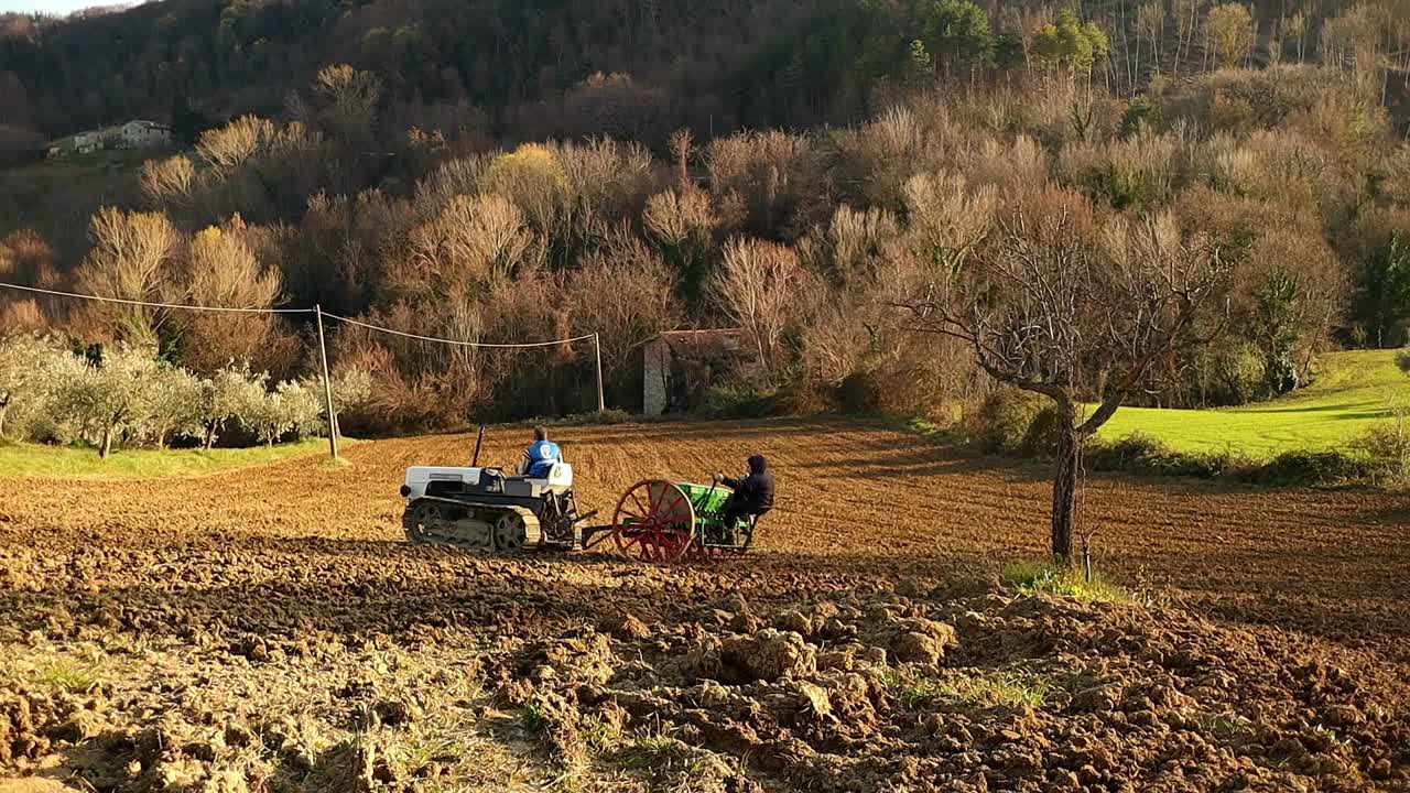 farmers working the land with crawler tractor and seeder at sunset, long shot