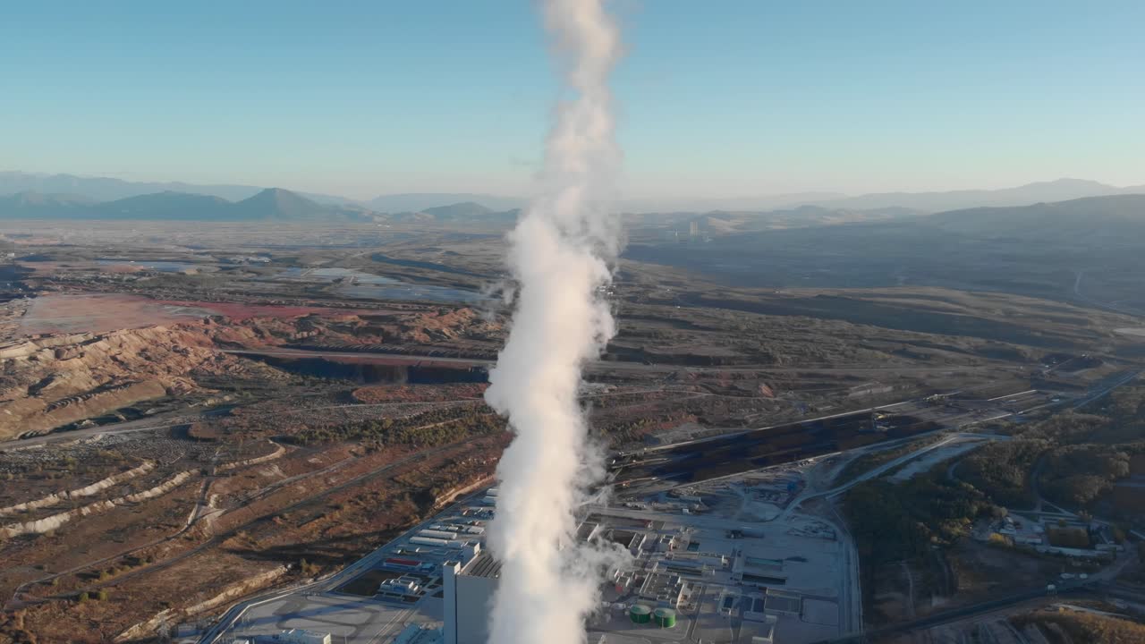Drone che segue fumo e vapore da una centrale termoelettrica a carbone, ripresa in picchiata al tramonto in un paesaggio rurale.