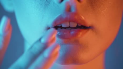 primer plano extremo de una mujer joven mientras ella seductoramente acaricia sus labios con su dedo y los presiona juntos con contrastes azul naranja en su cara