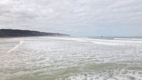 Low-aerial-shot-above-multiple-waves-crashing-on-the-shoreline-of-Muriwai-Beach,-New-Zealand