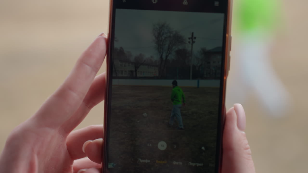 primer plano de la pantalla de un móvil en el que se ve a un niño con una chaqueta verde en un campo abierto; se aprecia la interfaz de grabación y el botón rojo; luz cálida de exteriores y edificio distante al fondo; captura íntima de un juego