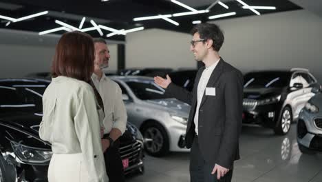 A-confident-male-assistant-in-a-car-dealership-tells-the-client-his-family-his-wife-and-little-son-about-what-cars-are-in-the-showroom-and-about-the-cars.-Confident-young-male-brunette-assistant-in-a-business-suit-talks-about-family-about-cars-in-a-car-showroom