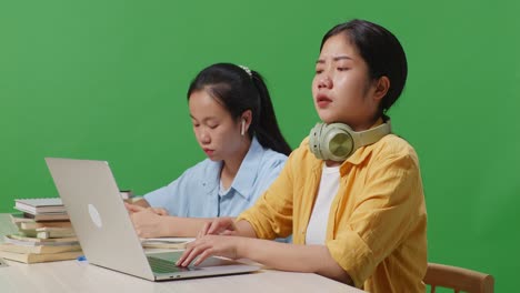 close up of asian woman student typing on a laptop and stretching while sitting with her friend on the table in the green screen background classroom