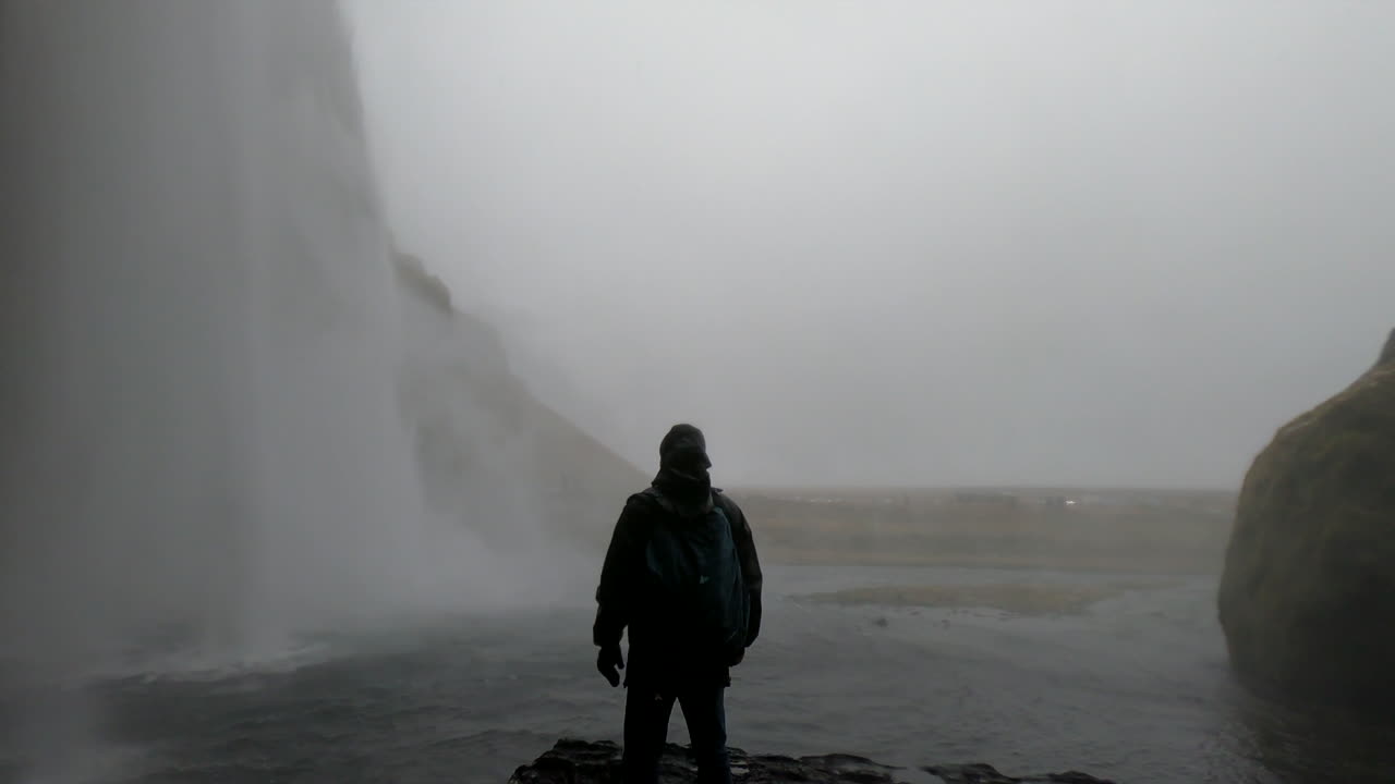 toma cinematográfica detrás de la cascada islandesa seljalandsfoss y donde un hombre admira la belleza y la fuerza del agua que cae